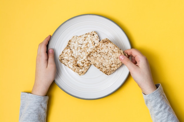 건강한 식생활. 귀리, 밀, 아마 및 참깨로 만든 Crispbread는 접시에 노란색 배경에 손을 얹습니다. 평면도