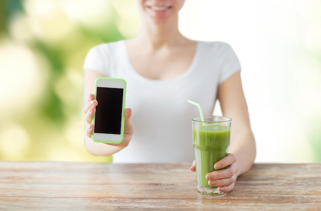 healthy eating, diet, detox, technology and people concept - close up of woman with smartphone green juice sitting at wooden table over green natural background