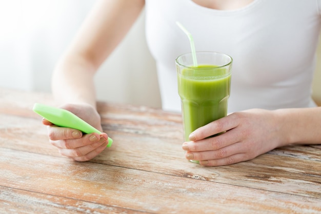 healthy eating, diet, detox, technology and people concept - close up of woman with smartphone green juice sitting at wooden table