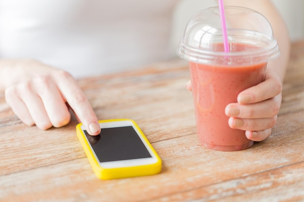 healthy eating, diet, technology and people concept - close up of woman with smartphone and cup of smoothie sitting at table