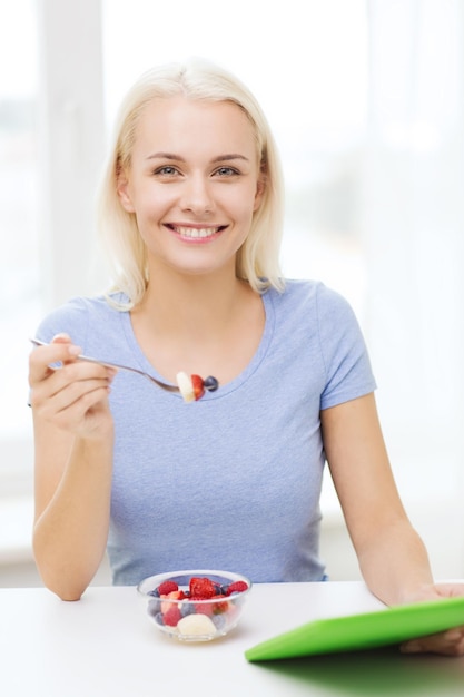 здоровое питание, диета и концепция людей - улыбающаяся молодая женщина с планшетным компьютером ест фруктовый салат дома