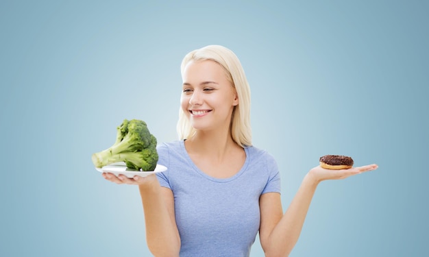 healthy eating, junk food, diet and choice people concept - smiling woman choosing between broccoli and donut over blue background