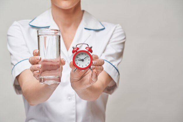 Healthy eating or lifestyle concept - female woman doctor holding and a glass of clear fresh waterand small red alarm clock