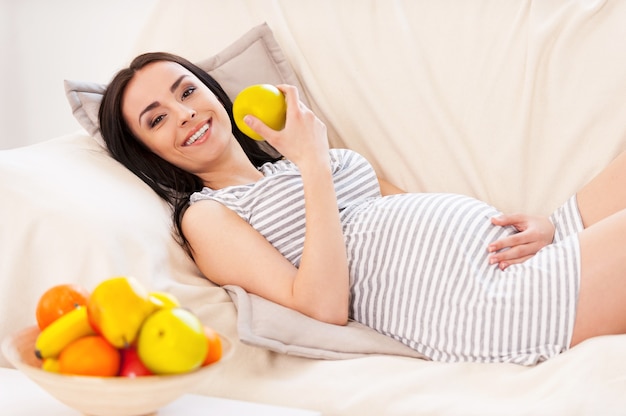 私と私の赤ちゃんのための健康的な食事。フルーツサラダを食べてソファに横になって笑っている美しい妊婦