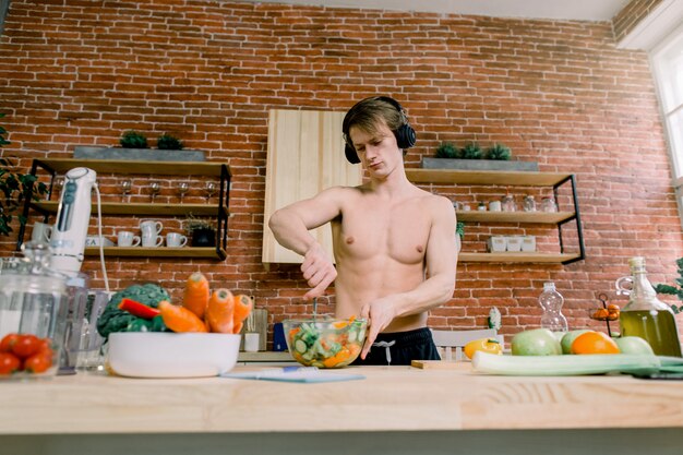 Healthy Eating And Nutrition Supplements. Handsome Male listening to music and dancing while preparing a salad Enjoying Sports Drink In Kitchen. Bodybuilding Concept