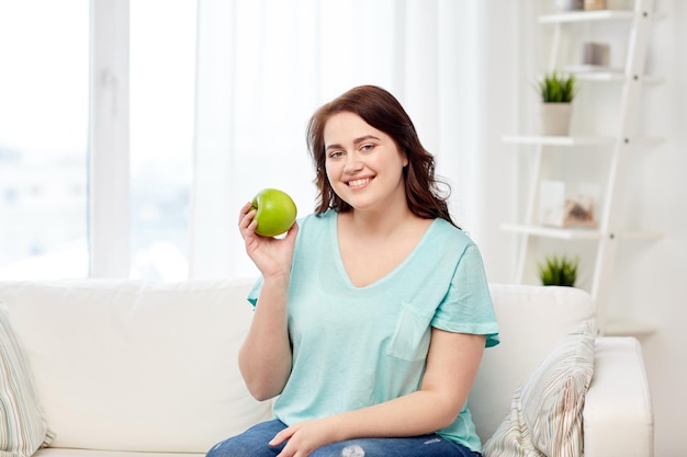 healthy eating, organic food, fruits, diet and people concept - happy young plus size woman eating green apple at home