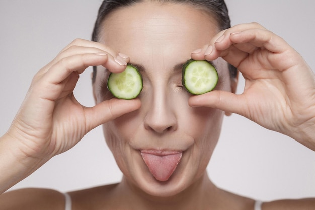 Healthy eyes. Portrait of beautiful middle aged woman with bare shoulders covering eyes with slice of cucumber and tongue out, isolated on grey background. Copy space, indoor, studio shot,isolated