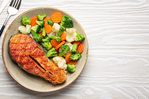 Healthy fish dinner: grilled salmon fish steak with vegetables salad on ceramic plate with fork and knife on white wooden background from above with copy space, clean and diet eating