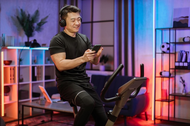 Healthy fit smiling asian man training at home on exercise static bike during workout holding phone