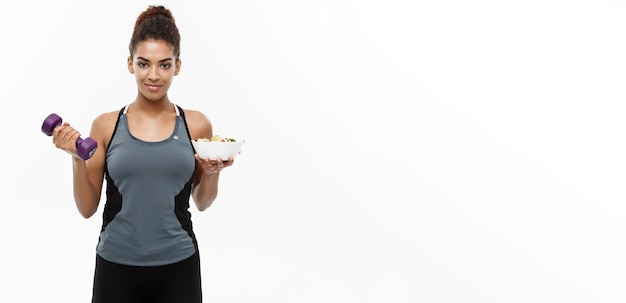 Healthy and Fitness concept Beautiful sporty African American on diet holding dumbbell and fresh salad on hands Isolated on white studio background