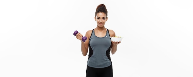 Healthy and Fitness concept Beautiful sporty African American on diet holding dumbbell and fresh salad on hands Isolated on white studio background