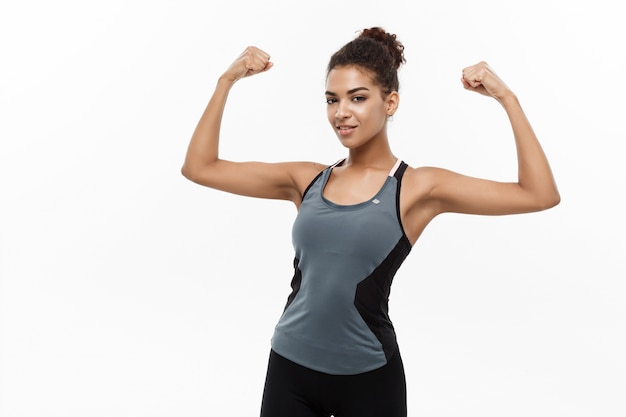 Healthy and Fitness concept - Portrait of young beautiful African American showing her strong muscle with confident cheerful facial expression. Isolated on white studio background.