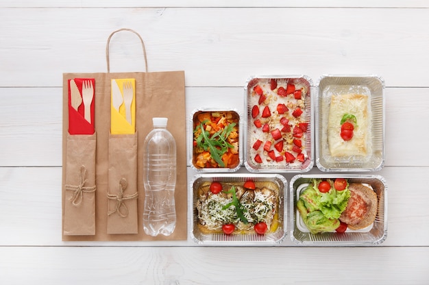 Healthy food delivery, daily meals and snacks. Nutrition, vegetables, meat, water bottle and fruits in foil boxes and brown paper package. Top view, flat lay at white wood with copy space