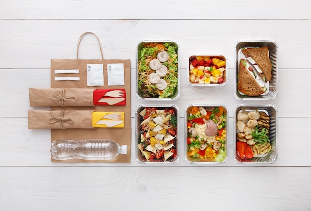 Healthy food delivery. Take away food. vegetables, meat and fruits in foil boxes, cutlery, water and brown paper package. Top view, flat lay at white wood with copy space