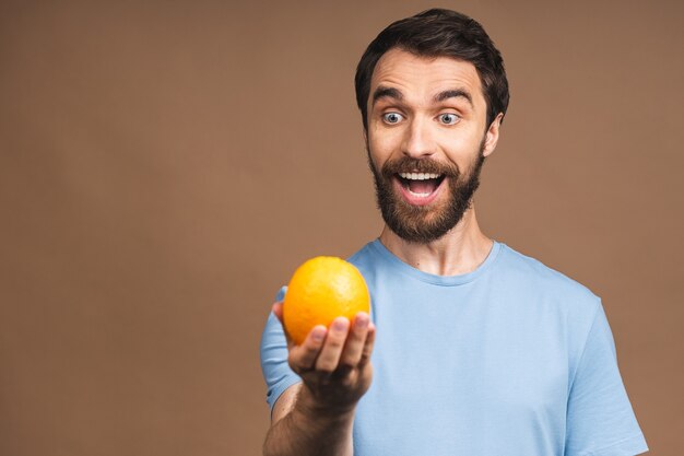 Healthy food and diet concept. Portrait of young bearded smiling man holding an orange fruit isolated on beige background.