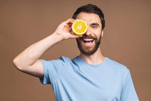 Healthy food and diet concept. Portrait of young bearded smiling man holding an orange fruit isolated on beige background.