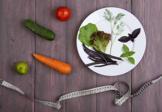 Healthy food and diet vegetarian concept - measure tape, white plate with leaf of salad, cucumber, tomato and other vegetables on wooden table