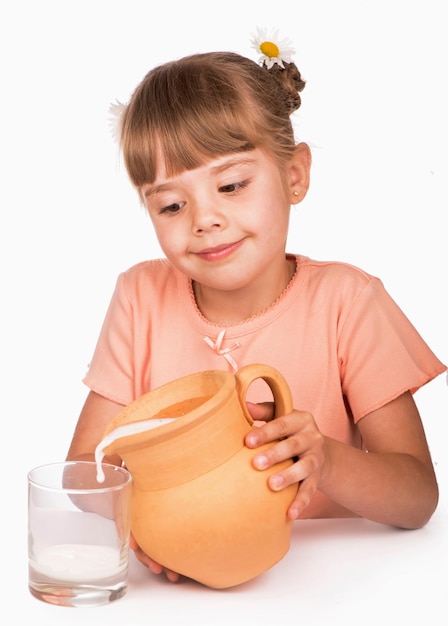 Healthy food A little girl in an orange Tshirt and flowers in her hair pours fresh milk from a jug on a white background