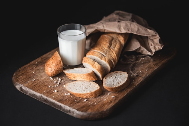 Здоровая пища. Батон деревенского хлеба с двумя отрезанными кусочками лежит на деревянной разделочной доске и стакане свежего молока. Темный фон. Горизонтальный снимок