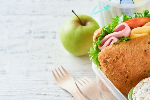 Healthy food for school lunch concept Healthy bread meat sandwich with cheese apple fresh cucumber carrots nuts in container water and yogurt on kitchen background Back to school concept