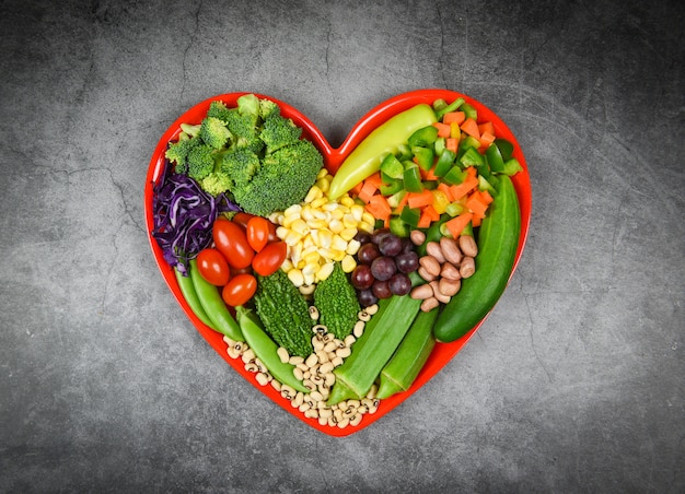 Selezione di alimenti sani mangiar pulito per la vita del cuore colesterolo dieta salute insalata fresca frutta e verdure verdi miste vari fagioli noci grano sul piatto cuore rosso per cibo sano cuoco vegano