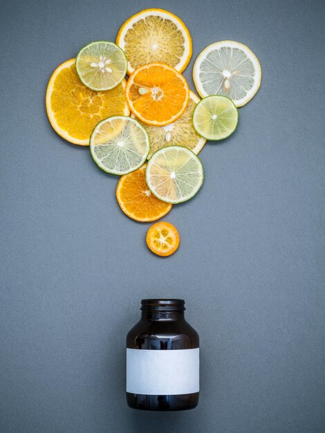 Фото Концепция здоровой пищи и лекарств бутылка с витамином с и различными цитрусовыми фруктами цитрусовые нарезки лимон, апельсин и лимон на сером фоне