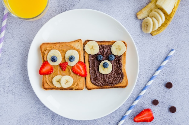 Healthy funny face sandwiches for kids. Animal faces toast with peanut and hazelnat chocolate butter, banana, strawberry and blueberry on a white plate with orange juice. Close up, Top view.