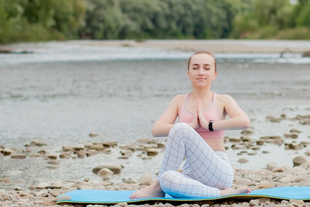 Healthy girl relaxing while meditating and doing yoga exercise in the beautiful nature on the bank of the river.