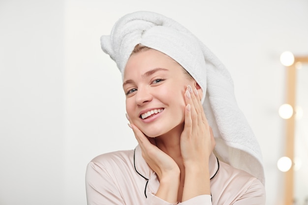 Healthy girl with toothy smile touching her clean hydrated face while enjoying its softness and radiance