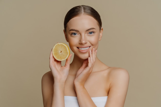 Healthy glad young woman poses with bare shoulders indoor wrapped in bath towel holds slice of lemon