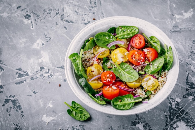 Здоровый салат из зеленой миски со шпинатом, киноа, желтыми и красными помидорами, луком и семенами на сером фоне