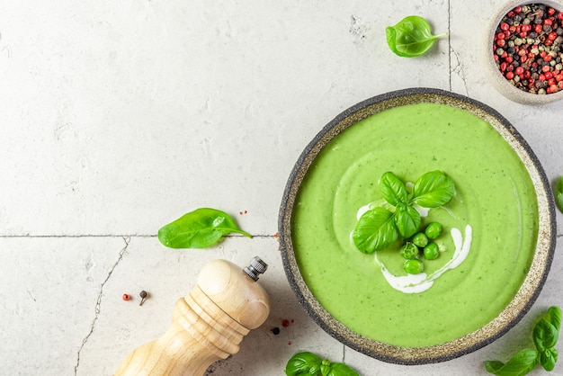 Healthy green cream pea and broccoli soup with basil in a bowl over light stone surface