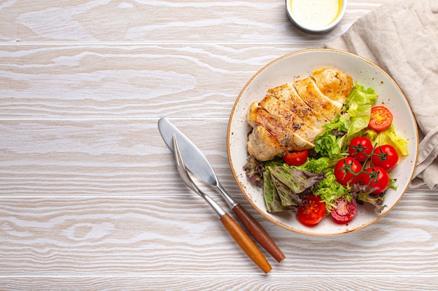 Healthy green vegetable salad with grilled chicken breast fillet on ceramic plate with olive oil on the side on white wooden kitchen table top view flat lay, diet food concept with space for text