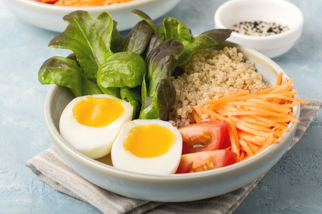 Healthy green vegetarian buddha bowl lunch with eggs, quinoa, carrots,tomato, lettuce leaves, on a blue background. close up