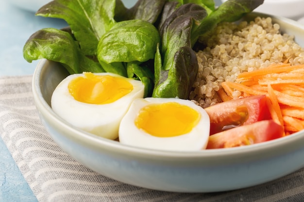 Healthy green vegetarian buddha bowl lunch with eggs, quinoa, carrots,tomato, lettuce leaves, on a blue background.