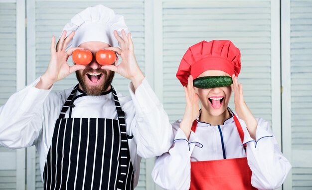 Foto dieta sana e felice maestro cuoco e aiutante che giocano con le verdure in cucina cuoco e cameriera di cucina che tengono pomodori e cetrioli agli occhi una coppia di cuochi che si divertono con cibo naturale in ristorante