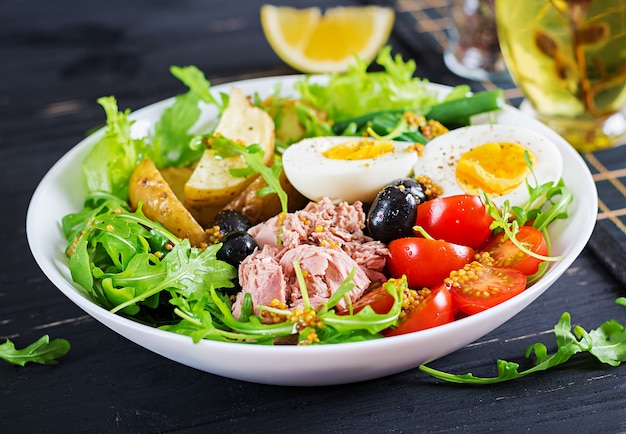 Здоровый сытный салат из тунца, фасоль, помидоры, яйца, картофель, маслины крупным планом в миску на столе. Салат Нисуаз. Французская кухня.