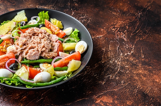 Healthy hearty salad with tuna, green beans, tomatoes, eggs, potatoes and black olives in a plate. Dark background.