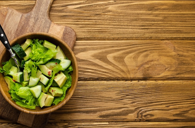 건강한 홈메이드 아보카도, 오이, 녹색 잎 샐러드는 나무로 된 소박한 갈색 배경의 커팅 보드에 있는 나무 그릇에 있습니다. 채식 음식, 다이어트를 위한 건강한 간식, 깨끗한 식사. 상위 뷰, 복사 공간