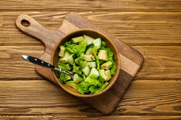건강한 홈메이드 아보카도, 오이, 녹색 잎 샐러드는 나무로 된 소박한 갈색 배경의 커팅 보드에 있는 나무 그릇에 있습니다. 채식 음식, 다이어트를 위한 건강한 간식, 깨끗한 식사. 평면도