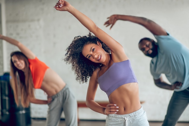 Healthy lifestyle. Cute young slim girl with dark curly hair doing warm-up with friends indoors