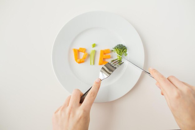 healthy lifestyle, diet, vegetarian food and people concept - close up of woman with fork and knife eating vegetable letters on plate