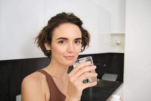 Photo healthy lifestyle and female wellbeing young beautiful woman standing in kitchen and drinking glass