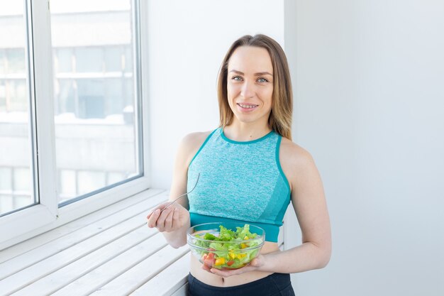 Stile di vita sano, fitness e concetto di dieta: insalata dietetica e donna magra in abbigliamento sportivo vicino alla finestra