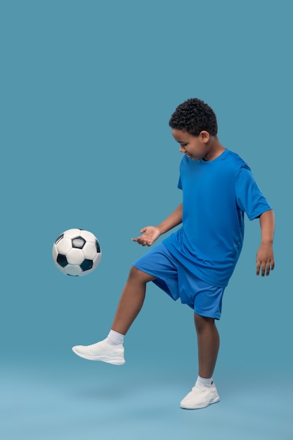 Healthy lifestyle. Interested involved african american elementary school boy in sportswear and sneakers playing soccer on blue background