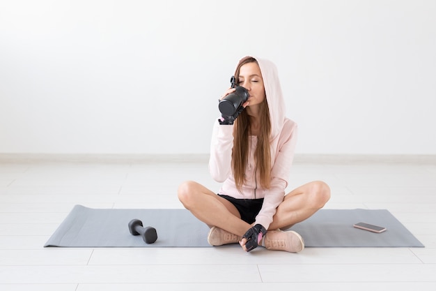 Здоровый образ жизни, люди и спортивная концепция - улыбается женщина, сидящая на коврике для йоги и питьевой воде после тяжелой тренировки.