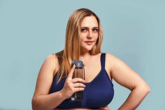 健康的な生活様式。水のボトルを保持し、青い壁に立ってカメラを見ているスポーツ服の若いプラスサイズの女性