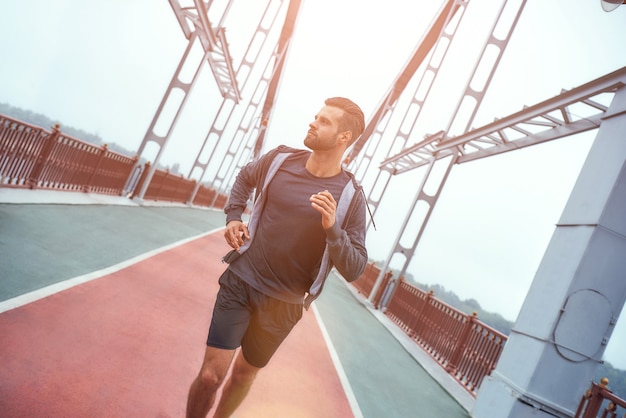 橋の上でジョギングして見ているスポーツ服の健康的なライフスタイルの若い都会のひげを生やした男