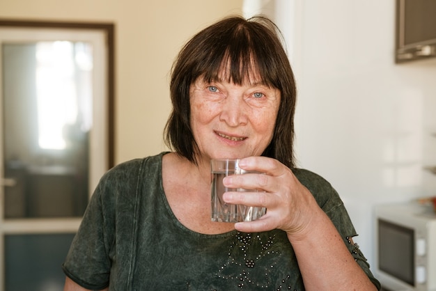 Healthy liquid caucasian mature woman drinks clear water from glass standing in the kitchen by the w...