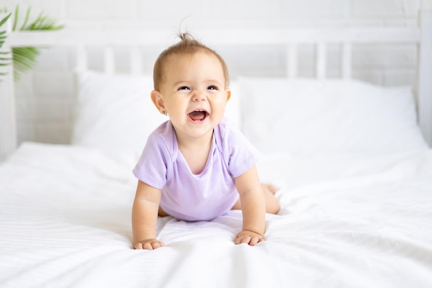 Healthy little cute girl child in a bodysuit crawls on the bed on white linens looks at the camera laughs The concept of baby products